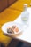 A white plate on a table holds a gourmet dish featuring colorful garnishes, grilled vegetables, and a delicate, lacy edible decoration. Next to the plate, there is a clear glass bottle with a cork stopper and a label displaying the word 'Anaë'. The background includes a soft, warm-toned cushioned seat.