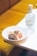A white plate on a table holds a gourmet dish featuring colorful garnishes, grilled vegetables, and a delicate, lacy edible decoration. Next to the plate, there is a clear glass bottle with a cork stopper and a label displaying the word 'Anaë'. The background includes a soft, warm-toned cushioned seat.