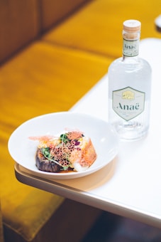 A white plate on a table holds a gourmet dish featuring colorful garnishes, grilled vegetables, and a delicate, lacy edible decoration. Next to the plate, there is a clear glass bottle with a cork stopper and a label displaying the word 'Ana&euml;'. The background includes a soft, warm-toned cushioned seat.