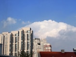 A cluster of tall, modern apartment buildings with numerous windows is set against a backdrop of fluffy, white clouds in a clear blue sky. In the foreground, a red-tiled roof of a smaller building is partially visible.