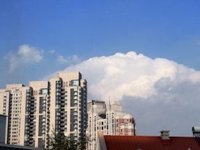 A cluster of tall, modern apartment buildings with numerous windows is set against a backdrop of fluffy, white clouds in a clear blue sky. In the foreground, a red-tiled roof of a smaller building is partially visible.