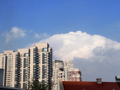 A cluster of tall, modern apartment buildings with numerous windows is set against a backdrop of fluffy, white clouds in a clear blue sky. In the foreground, a red-tiled roof of a smaller building is partially visible.
