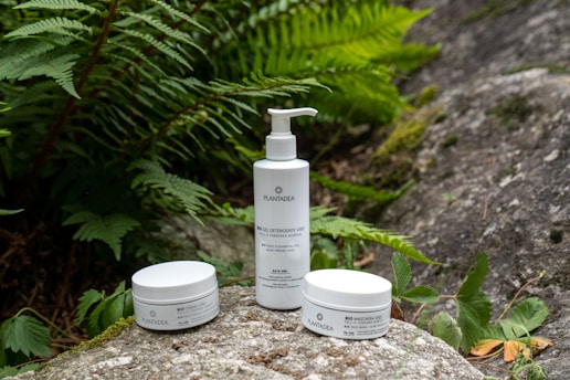 Three white cosmetic containers, including a pump bottle, are placed on a rock surrounded by lush green ferns and other natural foliage.