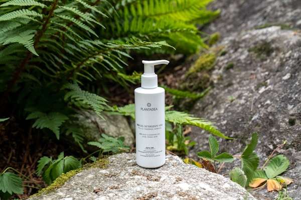 A bottle of skincare gel by Plantea is placed outdoors on a mossy rock. Surrounding the bottle, there are lush green ferns and small plants, contributing to a natural and organic aesthetic. The setting includes additional rocks, enhancing the earthy environment.