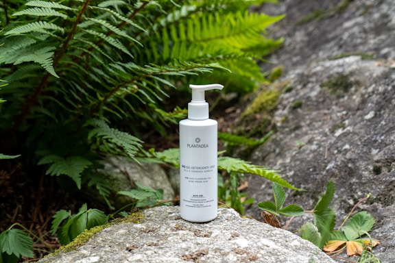 A white bottle of skincare product with a pump top is placed on a mossy rock surrounded by lush green ferns and leafy plants. The background consists of natural stone surfaces, creating a serene and earthy atmosphere.