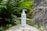 A white bottle of skincare product with a pump top is placed on a mossy rock surrounded by lush green ferns and leafy plants. The background consists of natural stone surfaces, creating a serene and earthy atmosphere.