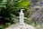 A white bottle of skincare product with a pump top is placed on a mossy rock surrounded by lush green ferns and leafy plants. The background consists of natural stone surfaces, creating a serene and earthy atmosphere.