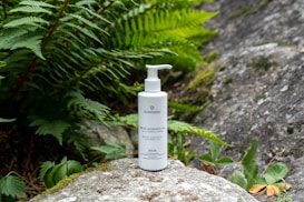 A white bottle of skincare product with a pump top is placed on a mossy rock surrounded by lush green ferns and leafy plants. The background consists of natural stone surfaces, creating a serene and earthy atmosphere.