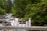 A collection of skincare products is displayed on a wooden railing with a forest stream in the background. The products include tubes and bottles in white packaging, positioned neatly in a row. Behind them, lush green trees and smooth rocks frame the gently flowing water of the stream.