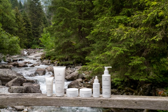 A collection of skincare products is displayed on a wooden railing with a forest stream in the background. The products include tubes and bottles in white packaging, positioned neatly in a row. Behind them, lush green trees and smooth rocks frame the gently flowing water of the stream.