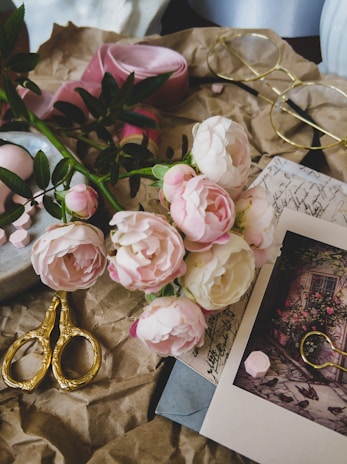 A collection of vintage-style objects is arranged on crumpled brown paper. There are pink and white peonies, a pink ribbon, ornate gold scissors, round glasses, and a photograph of a floral scene overlaid on handwritten letters.