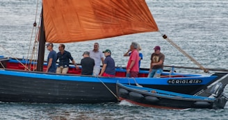 A group of people is sailing on a wooden boat with a vibrant red-orange sail. The boat features blue and black accents, and several men are standing or seated, engaging in conversation as it moves across a body of water.
