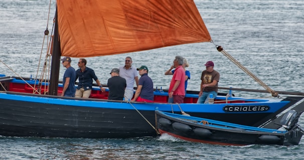 A group of people is sailing on a wooden boat with a vibrant red-orange sail. The boat features blue and black accents, and several men are standing or seated, engaging in conversation as it moves across a body of water.
