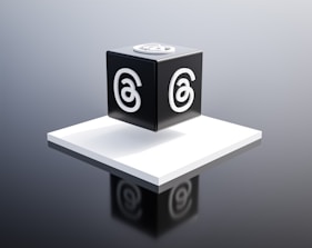A black cube with a stylized letter 'a' in white on each visible face is placed on a raised white platform. The background is a gradient of gray, giving the scene a modern and minimalist aesthetic.