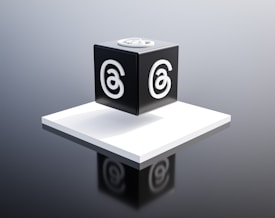 A black cube with a stylized letter 'a' in white on each visible face is placed on a raised white platform. The background is a gradient of gray, giving the scene a modern and minimalist aesthetic.