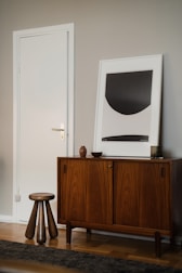 A modern interior scene featuring a mid-century style wooden cabinet with stylish grain patterns. A simple, abstract art piece in a frame rests on top of the cabinet. Beside the furniture, a short wooden stool with three legs adds a touch of minimalist design. The walls are painted in a neutral tone, complementing the wooden textures.