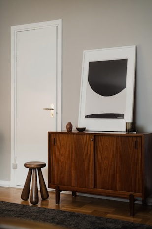 A modern interior scene featuring a mid-century style wooden cabinet with stylish grain patterns. A simple, abstract art piece in a frame rests on top of the cabinet. Beside the furniture, a short wooden stool with three legs adds a touch of minimalist design. The walls are painted in a neutral tone, complementing the wooden textures.