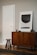 A modern interior scene featuring a mid-century style wooden cabinet with stylish grain patterns. A simple, abstract art piece in a frame rests on top of the cabinet. Beside the furniture, a short wooden stool with three legs adds a touch of minimalist design. The walls are painted in a neutral tone, complementing the wooden textures.
