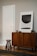A modern interior scene featuring a mid-century style wooden cabinet with stylish grain patterns. A simple, abstract art piece in a frame rests on top of the cabinet. Beside the furniture, a short wooden stool with three legs adds a touch of minimalist design. The walls are painted in a neutral tone, complementing the wooden textures.