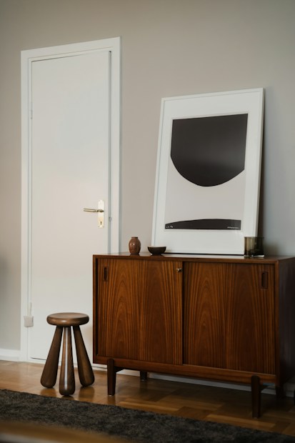 A modern interior scene featuring a mid-century style wooden cabinet with stylish grain patterns. A simple, abstract art piece in a frame rests on top of the cabinet. Beside the furniture, a short wooden stool with three legs adds a touch of minimalist design. The walls are painted in a neutral tone, complementing the wooden textures.
