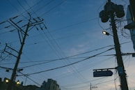 A network of utility poles and power lines against an evening sky, with a mix of clouds and fading daylight. Some streetlights are on, casting a warm glow. Urban buildings are partially visible at the bottom.