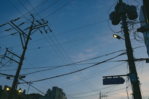 A network of utility poles and power lines against an evening sky, with a mix of clouds and fading daylight. Some streetlights are on, casting a warm glow. Urban buildings are partially visible at the bottom.