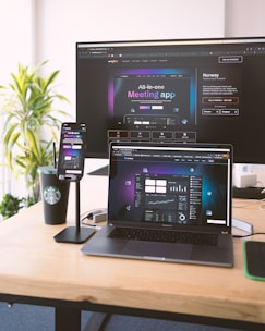Several electronic devices are placed on a wooden desk, including a large monitor, a laptop, and a smartphone mounted on a stand. All devices display a webpage for an all-in-one meeting app. There is also a Starbucks cup and some office supplies visible on the desk. A green plant is in the background, adding a touch of nature to the workspace.