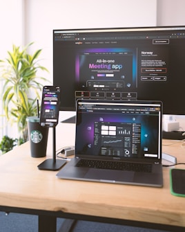 Several electronic devices are placed on a wooden desk, including a large monitor, a laptop, and a smartphone mounted on a stand. All devices display a webpage for an all-in-one meeting app. There is also a Starbucks cup and some office supplies visible on the desk. A green plant is in the background, adding a touch of nature to the workspace.
