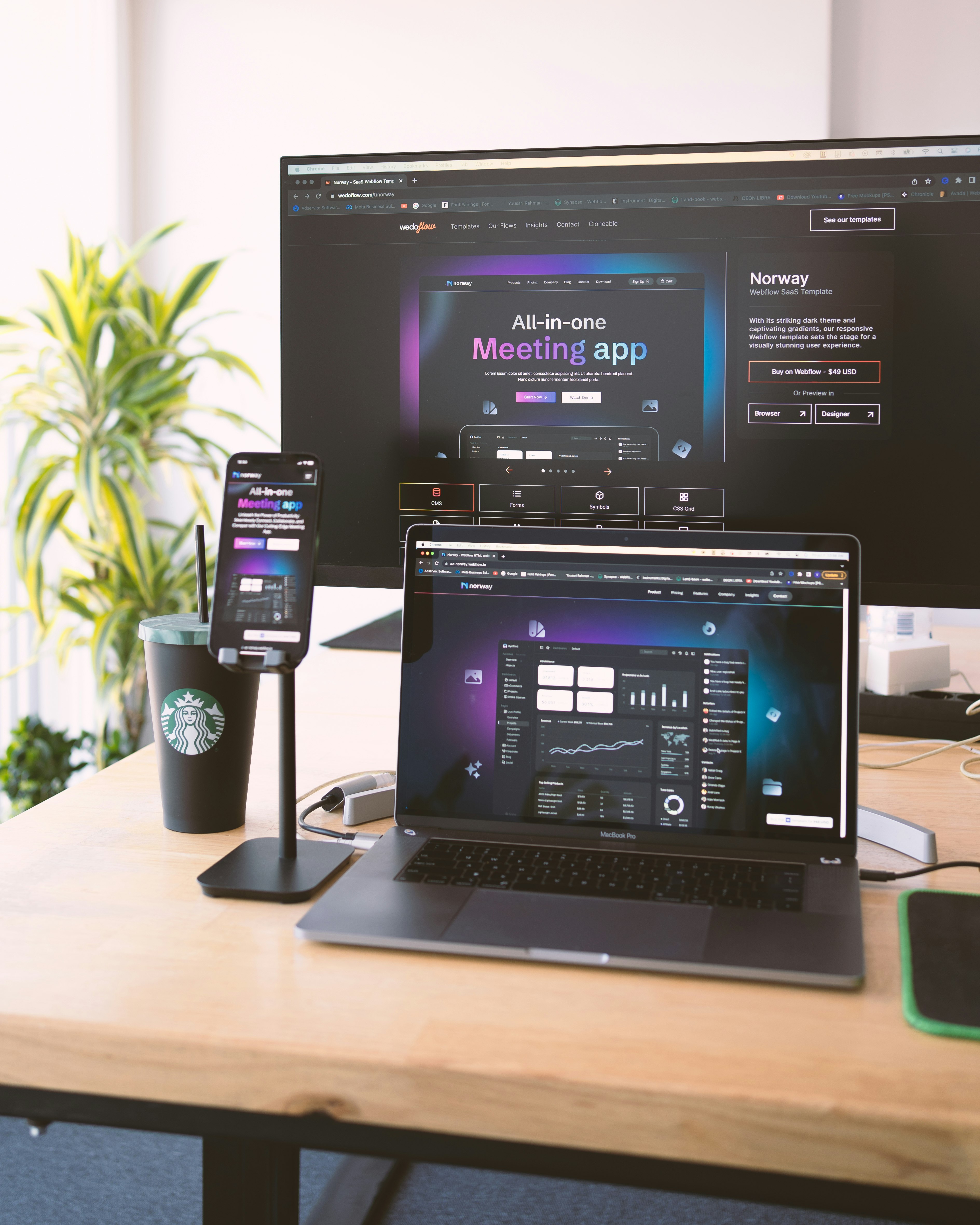 Several electronic devices are placed on a wooden desk, including a large monitor, a laptop, and a smartphone mounted on a stand. All devices display a webpage for an all-in-one meeting app. There is also a Starbucks cup and some office supplies visible on the desk. A green plant is in the background, adding a touch of nature to the workspace.