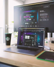 A laptop on a wooden desk displays a web application interface featuring graphs and statistics. Beside it, a large monitor shows a webpage promoting an all-in-one meeting app. A cup with a recognizable green and white logo sits on the desk.