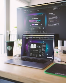 A laptop on a wooden desk displays a web application interface featuring graphs and statistics. Beside it, a large monitor shows a webpage promoting an all-in-one meeting app. A cup with a recognizable green and white logo sits on the desk.