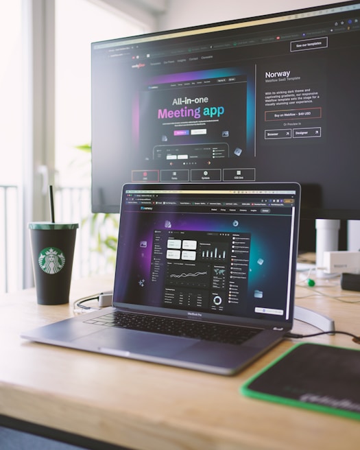 A laptop on a wooden desk displays a web application interface featuring graphs and statistics. Beside it, a large monitor shows a webpage promoting an all-in-one meeting app. A cup with a recognizable green and white logo sits on the desk.