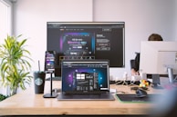 A modern workspace featuring multiple screens displaying a meeting app. A laptop, smartphone, and large monitor show similar interfaces, suggesting a professional setup. A Starbucks cup, Rubik's cube, and various desk items provide a casual atmosphere. A person is partially visible, working at a desk.