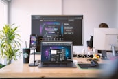 A modern workspace featuring multiple screens displaying a meeting app. A laptop, smartphone, and large monitor show similar interfaces, suggesting a professional setup. A Starbucks cup, Rubik's cube, and various desk items provide a casual atmosphere. A person is partially visible, working at a desk.