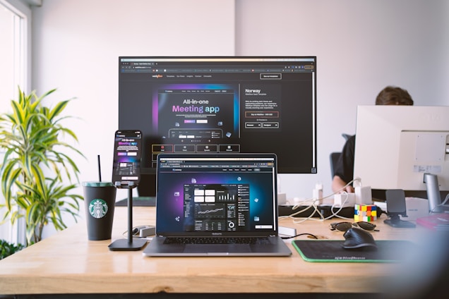 A modern workspace featuring multiple screens displaying a meeting app. A laptop, smartphone, and large monitor show similar interfaces, suggesting a professional setup. A Starbucks cup, Rubik's cube, and various desk items provide a casual atmosphere. A person is partially visible, working at a desk.