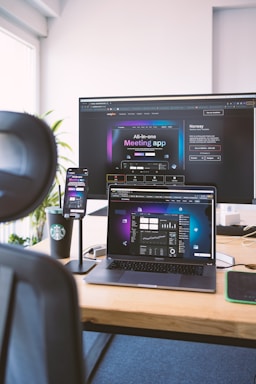 A workspace setup featuring an open laptop, smartphone, and large monitor displaying an interface for a meeting application. The environment includes a black chair visible in the foreground, a wooden desk, and a branded coffee cup. Light streams in from a window, illuminating a plant in the background.