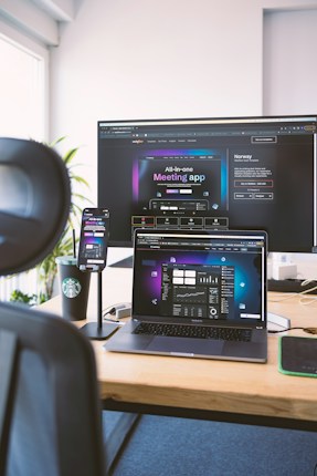 A workspace setup featuring an open laptop, smartphone, and large monitor displaying an interface for a meeting application. The environment includes a black chair visible in the foreground, a wooden desk, and a branded coffee cup. Light streams in from a window, illuminating a plant in the background.