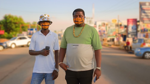 Two men stand on a city street. They are wearing face masks with intricate patterns. The man on the left wears a white shirt, blue jeans, and a bucket hat with designs. The man on the right is dressed in a green and beige shirt with accessories like a beaded necklace. Cars and buildings are visible in the blurred background, suggesting an urban setting.