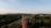 A brick chimney rises from a densely wooded area with distant hills under a clear sky. Small plants grow from the top of the chimney. A town with roads and scattered buildings is visible, with green trees prominent in the landscape.