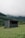 A small, simple shelter is situated in a lush, grassy meadow. In the background, there are densely forested hills partially shrouded in mist, giving the scene a serene and slightly mysterious atmosphere. The shelter appears modest with a flat roof and a combination of wood and concrete materials.
