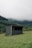 A small, simple shelter is situated in a lush, grassy meadow. In the background, there are densely forested hills partially shrouded in mist, giving the scene a serene and slightly mysterious atmosphere. The shelter appears modest with a flat roof and a combination of wood and concrete materials.