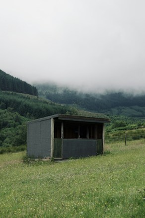 A small, simple shelter is situated in a lush, grassy meadow. In the background, there are densely forested hills partially shrouded in mist, giving the scene a serene and slightly mysterious atmosphere. The shelter appears modest with a flat roof and a combination of wood and concrete materials.