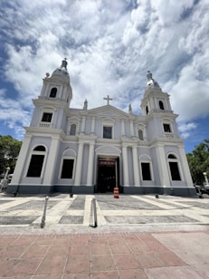 A large, white church with two towers and a central cross, set against a partly cloudy sky. The building features classical architectural elements such as columns and arches, and the facade is symmetrical with three entrances. The area in front of the church is paved and secured with chains, and an orange traffic cone is visible in front of the main entrance.