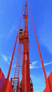 A tall red crane structure extends upward against a bright blue sky. The crane features a control cabin and a ladder for access, prominently highlighted in vibrant colors.