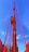 A tall red crane structure extends upward against a bright blue sky. The crane features a control cabin and a ladder for access, prominently highlighted in vibrant colors.