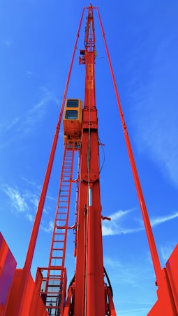A tall red crane structure extends upward against a bright blue sky. The crane features a control cabin and a ladder for access, prominently highlighted in vibrant colors.