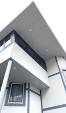 A modern building facade is shown with a combination of geometric shapes and clean lines. The architectural design features an overhanging upper floor with large windows and a supportive pillar. The structure has a minimalist design with light gray panels and blue accents around the windows.