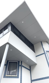 A modern building facade is shown with a combination of geometric shapes and clean lines. The architectural design features an overhanging upper floor with large windows and a supportive pillar. The structure has a minimalist design with light gray panels and blue accents around the windows.