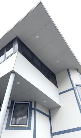 A modern building facade is shown with a combination of geometric shapes and clean lines. The architectural design features an overhanging upper floor with large windows and a supportive pillar. The structure has a minimalist design with light gray panels and blue accents around the windows.