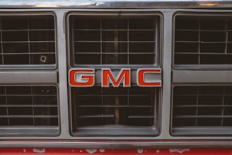 A close-up of a vehicle grille featuring the bold red letters 'GMC' prominently in the center. The grille is metallic with a grid pattern and shows some signs of wear.